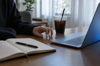 Main tapotant sur un bureau à côté d'une tasse de café froide, carnet ouvert et ordinateur portable allumé, décor de bureau lumineux et sobre